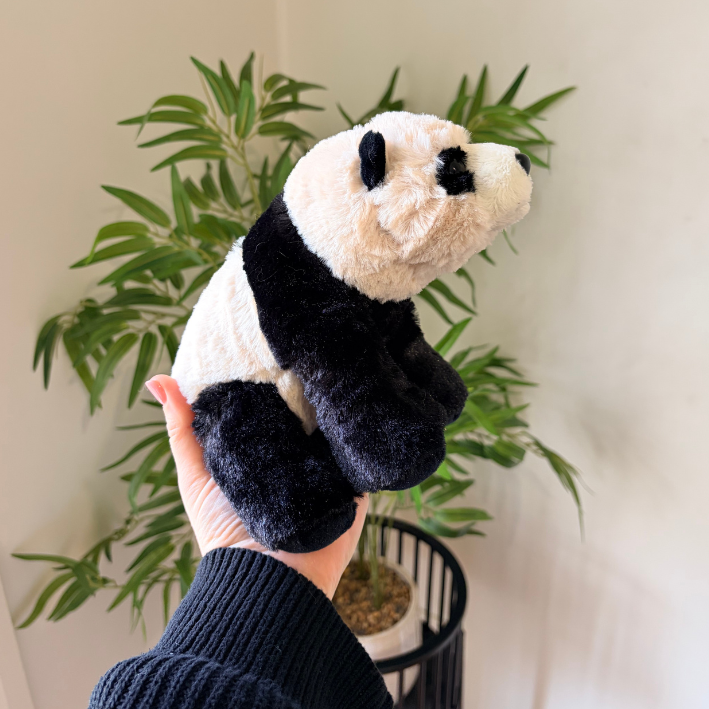 A plush panda toy being held up by a person's hand in front of a potted green plant against a light wall.