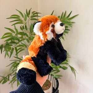 A plush red panda finger puppet being held by a hand in front of a green potted plant against a light beige wall.
