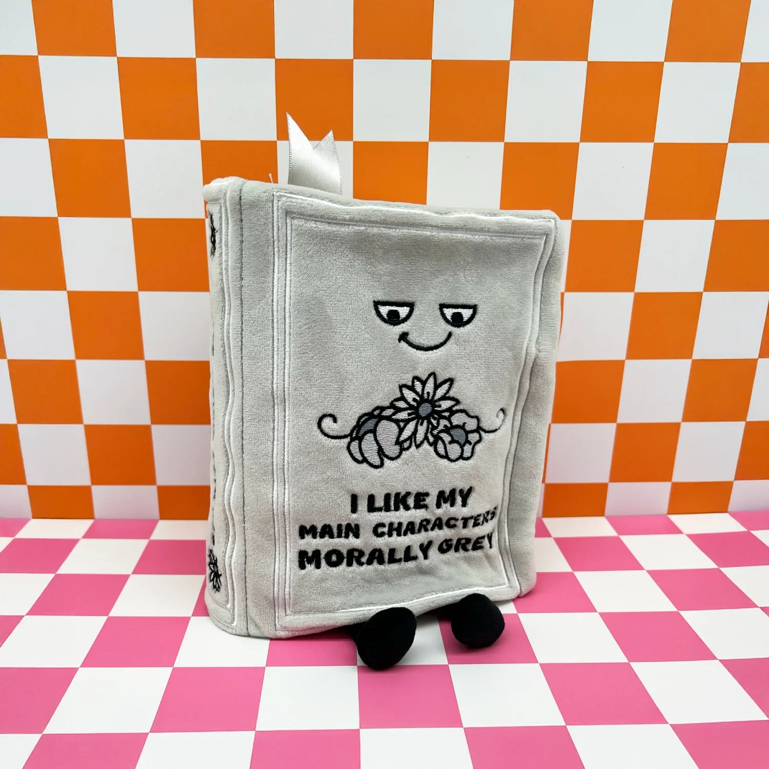 A plush gray book character toy with embroidered facial features, a flower motif, and the text 'I LIKE MY MAIN CHARACTER MORALLY GREW!' on the front, standing on a pink-and-white checkered surface against a matching orange-and-white checkered backdrop.