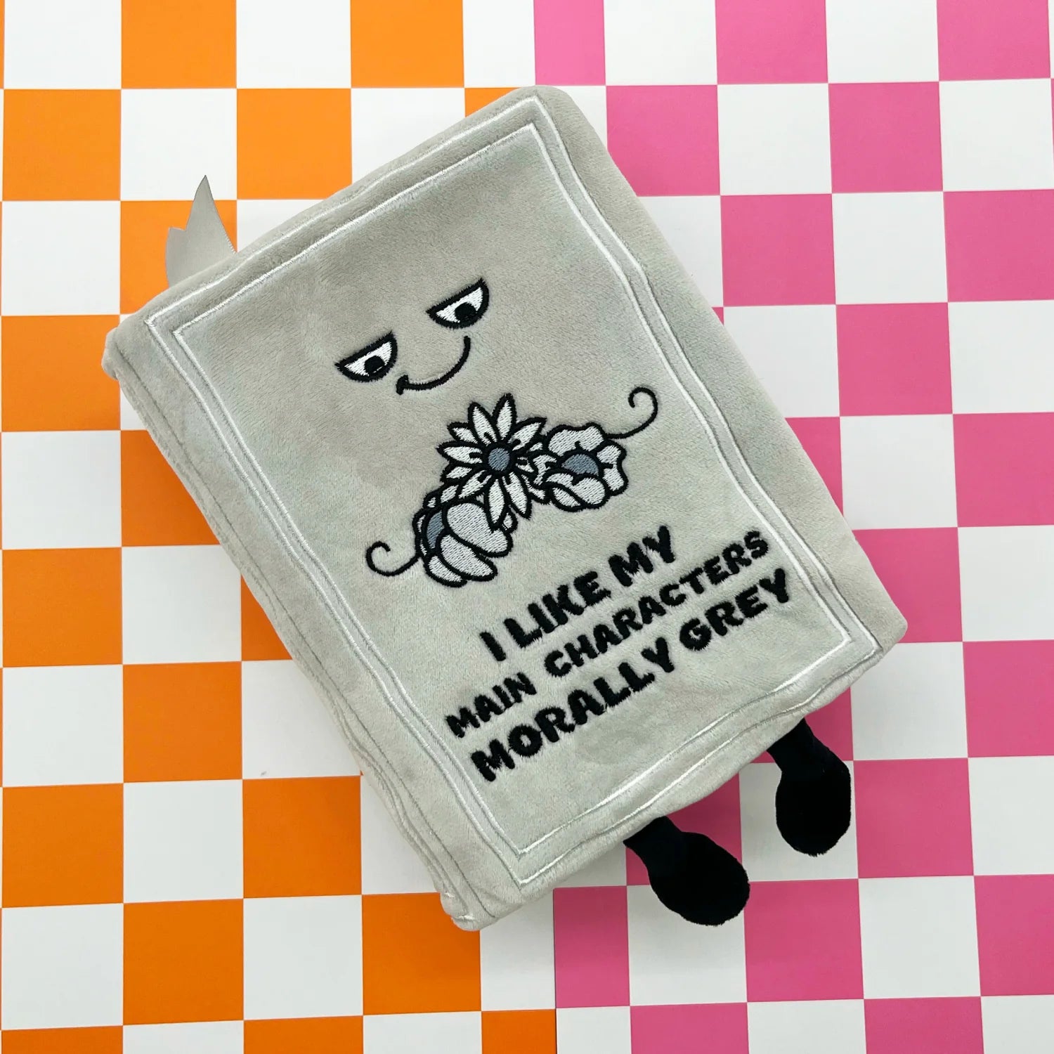 A plush grey book-shaped accessory with embroidered eyes, a flower, and the text 'I LIKE MY MAIN CHARACTERS MORALLY GREY' lies on a pink, white, and orange checkered background.
