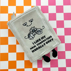 A plush grey book-shaped accessory with embroidered eyes, a flower, and the text 'I LIKE MY MAIN CHARACTERS MORALLY GREY' lies on a pink, white, and orange checkered background.