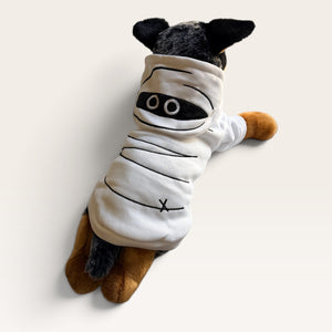 A small dog plushie wearing a white pet hoodie styled like a mummy, with black embroidered lines and circular eye details, photographed on a clean off-white background.