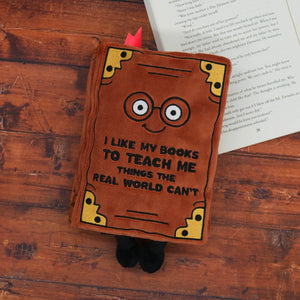 Plush book-shaped pillow with a face and text on a wooden surface