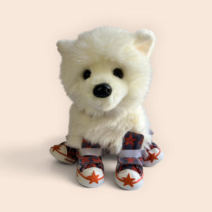 A white plush puppy toy wearing red and navy patterned booties with white star details, set against a solid pale peach background.
