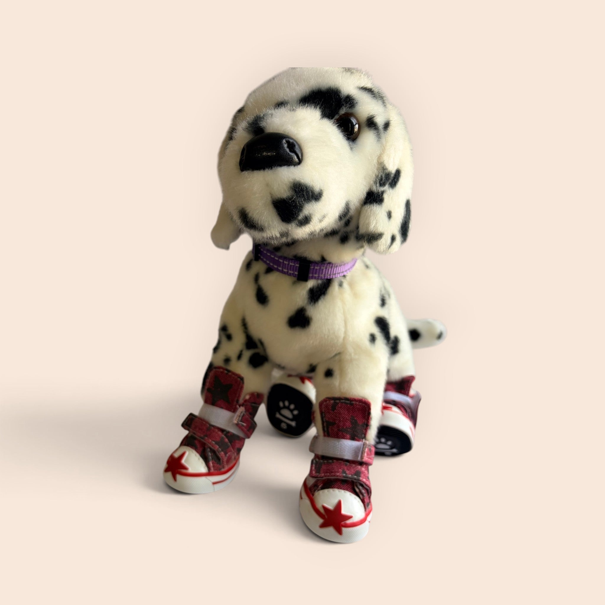 A plush Dalmatian dog toy wearing red and black plaid booties with white soles and red star details, set against a solid pale peach background.