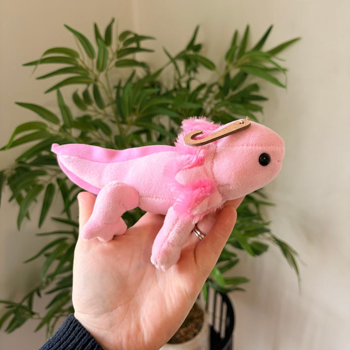 A small pink plush axolotl keychain or charm is held in a person's hand, featuring fluffy pink fabric, a small black bead eye, and a gold metal clip on its head. The background shows a blurred green potted plant against a light wall.