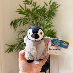 A small plush penguin toy held in a person's hand, with a blue hangtag reading Pocketkins. The background shows a blurred indoor plant against a light wall.