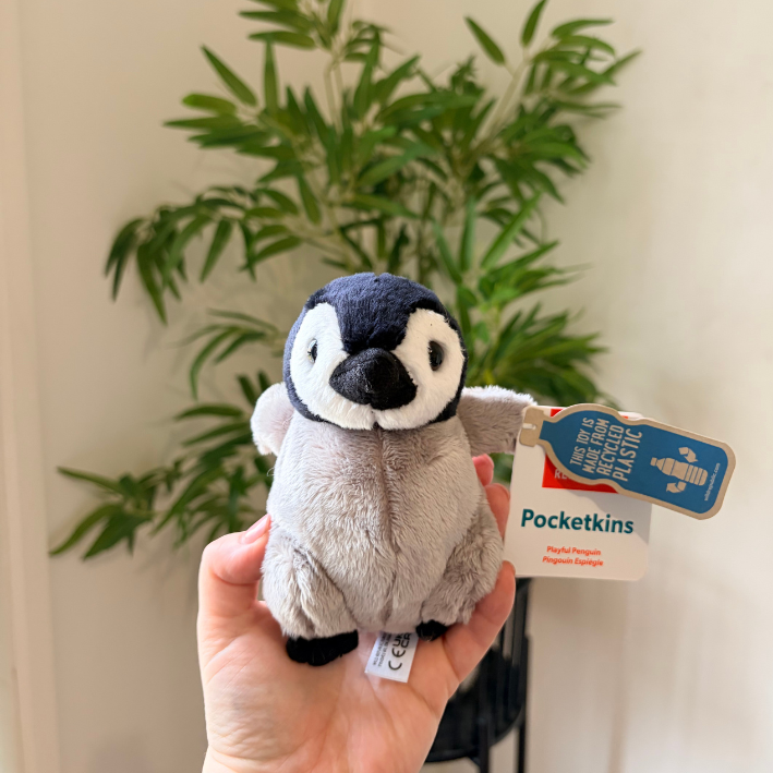 A small plush penguin toy held in a person's hand, with a blue hangtag reading Pocketkins. The background shows a blurred indoor plant against a light wall.