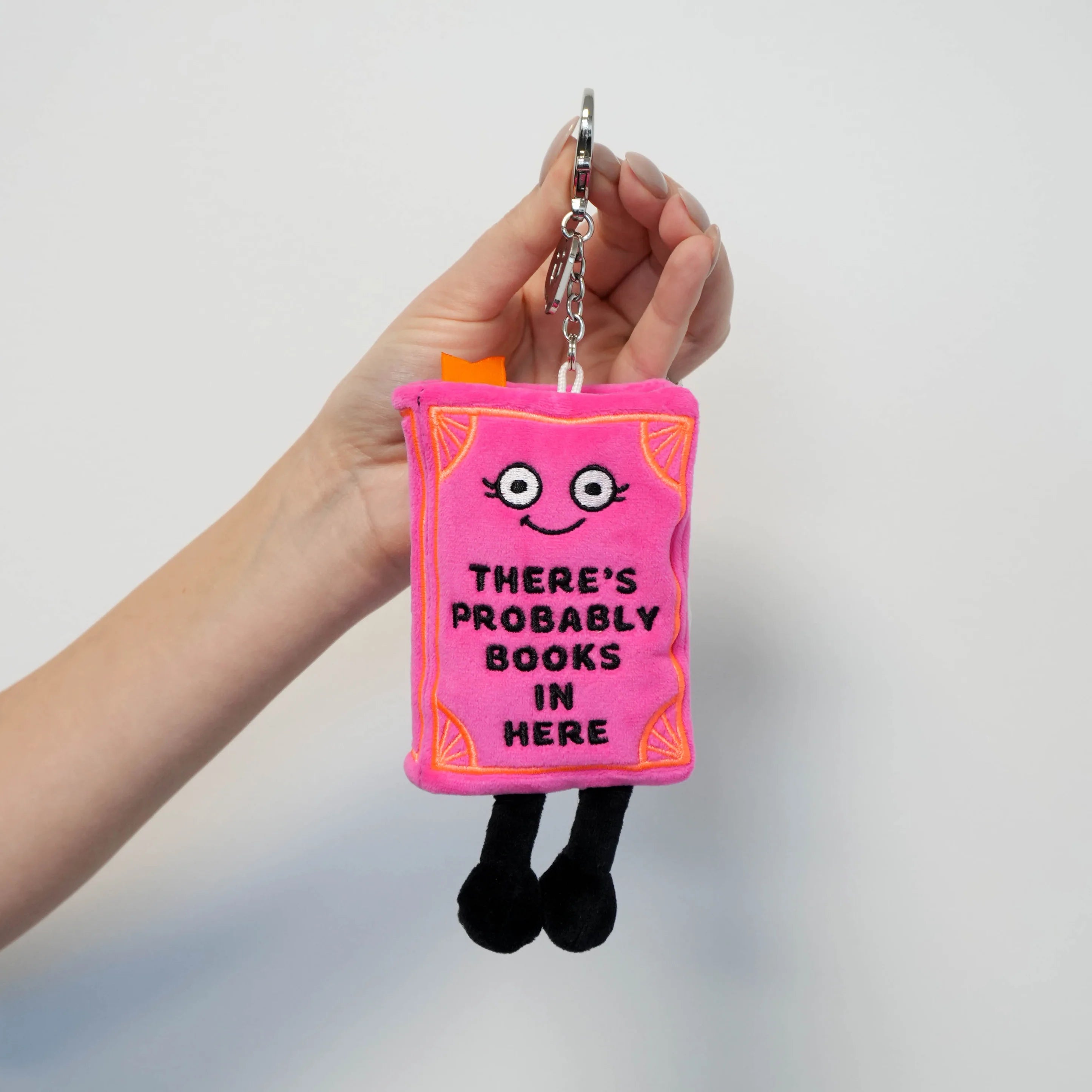 A hand holding a small plush book keychain charm. The charm is bright pink with black embroidered text reading 'THERE'S PROBABLY BOOKS IN HERE', a simple face, orange accents, and black legs.