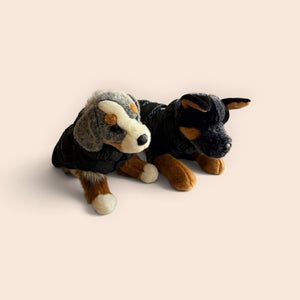Two plush dog toys wearing black jackets, posed side by side on a plain warm beige background.