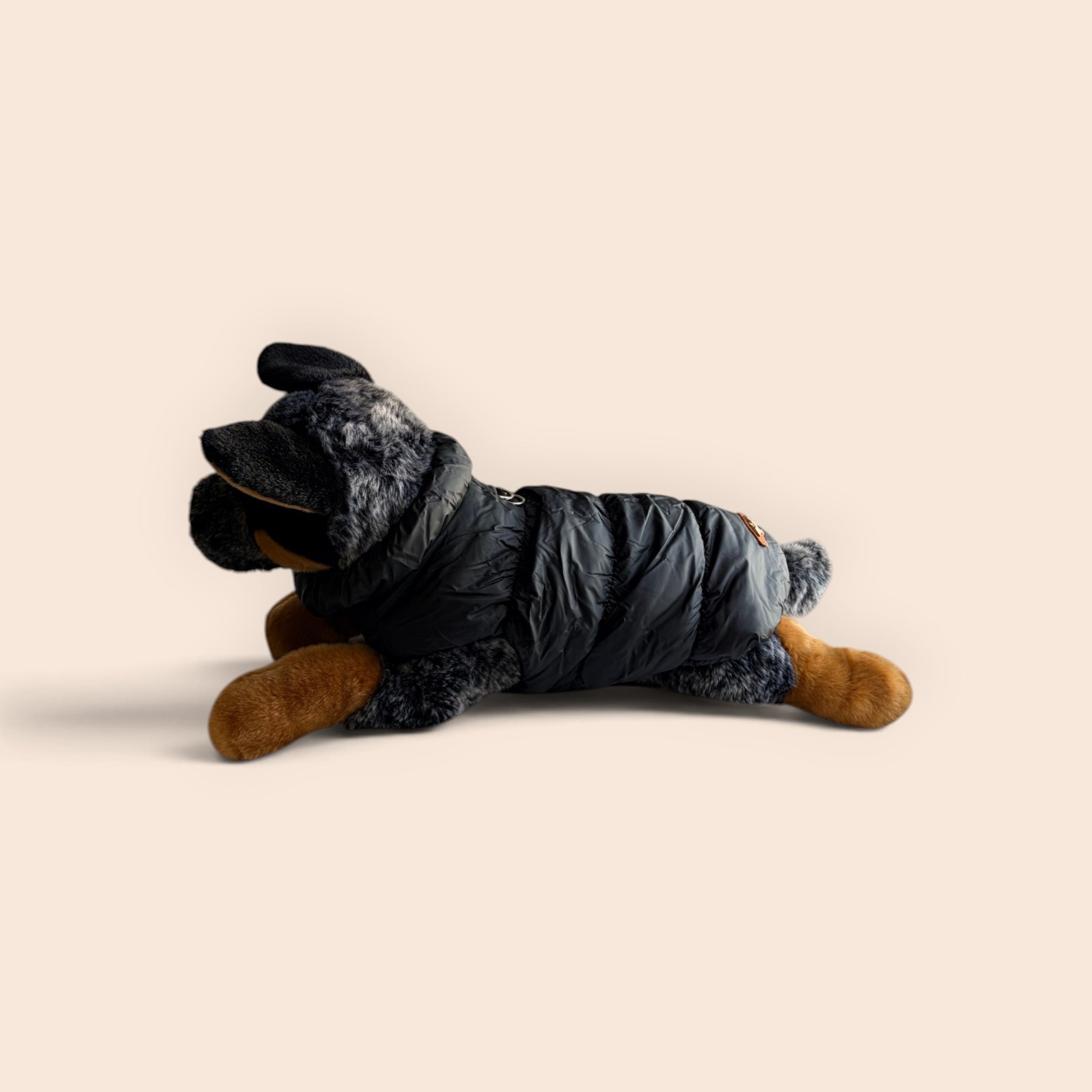 A plush dog toy wearing a black puffer jacket, posed on its side against a solid light beige background.