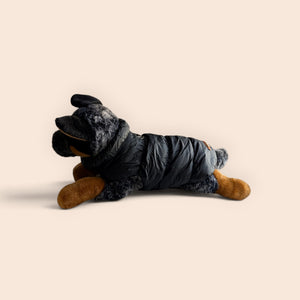 A plush dog toy wearing a black puffer jacket, posed on its side against a solid light beige background.