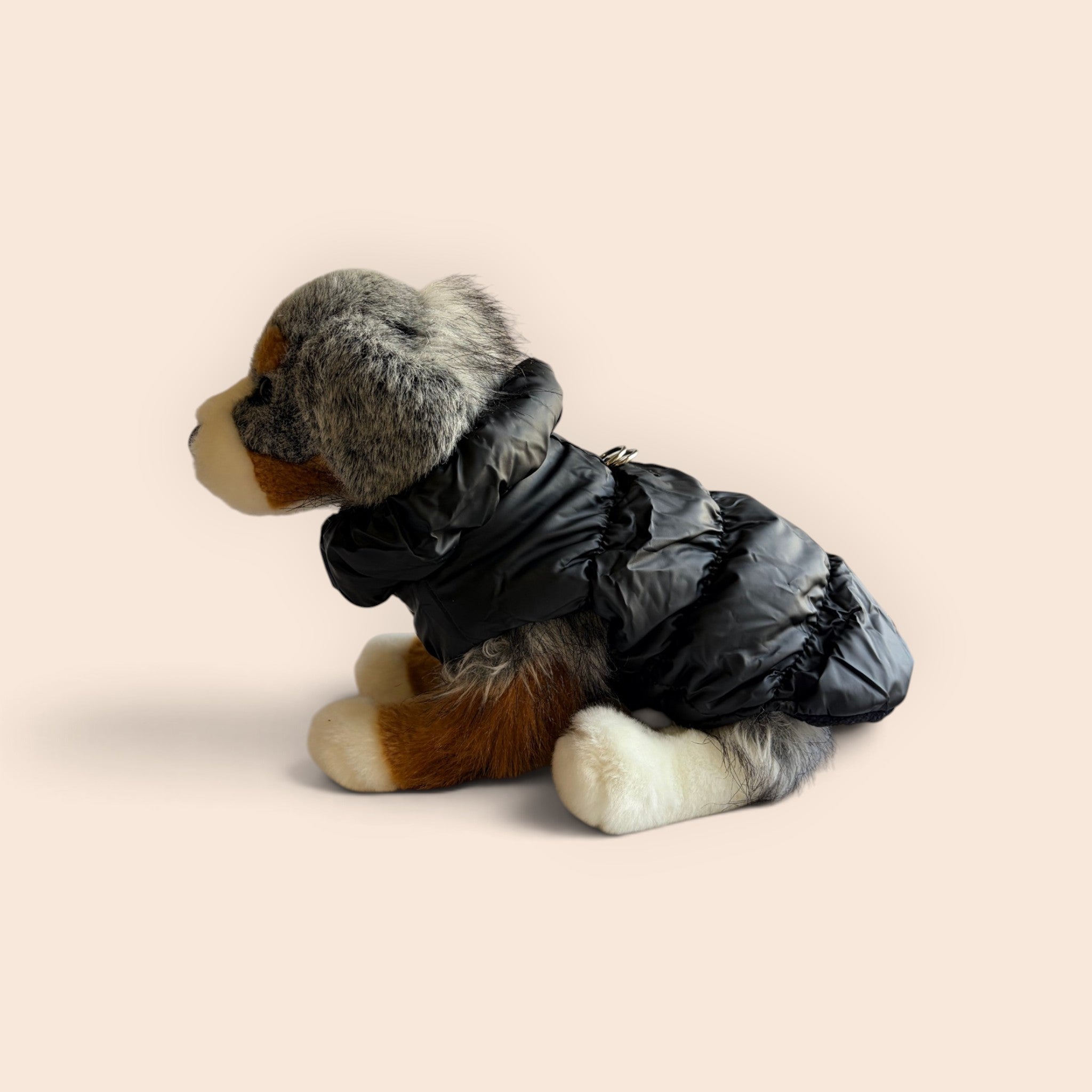 A small plush dog wearing a black puffer jacket, posed in profile against a solid pale peach background.