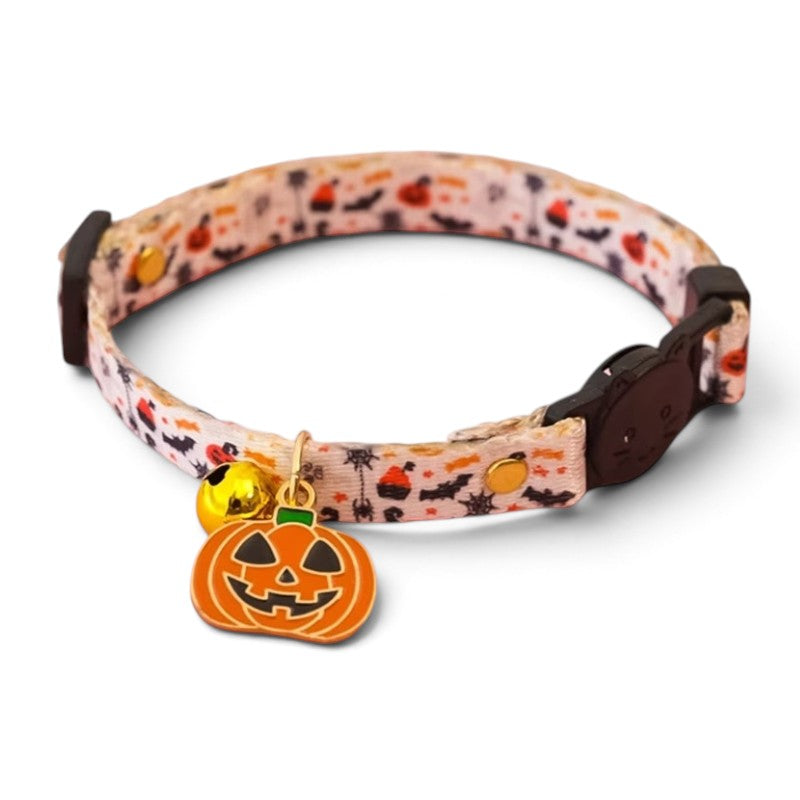 A dog collar with a white Halloween-themed pattern featuring pumpkins, bats, and ghosts, adorned with a dangling orange jack-o'-lantern charm and a small gold bell.