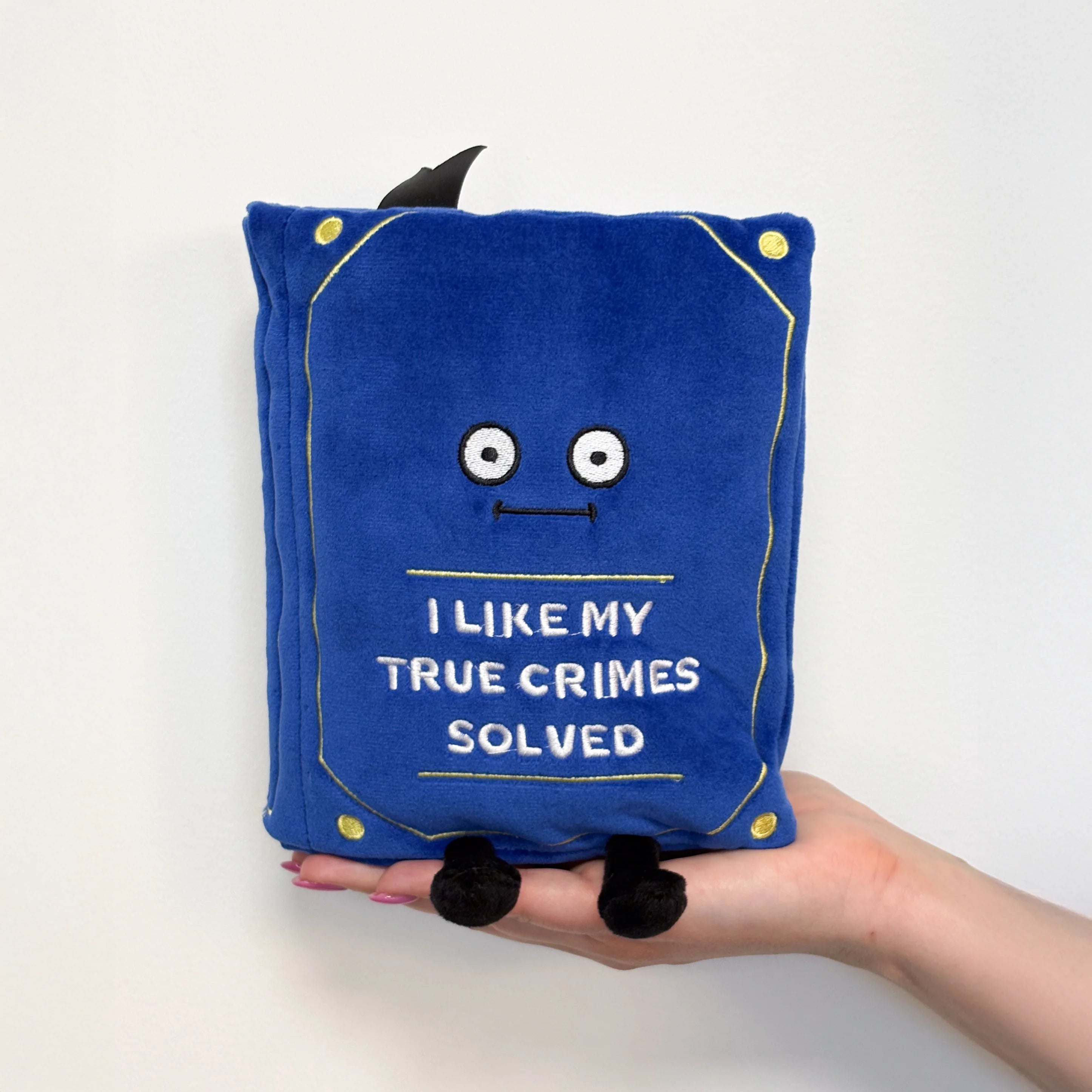 A blue plush book-shaped pillow with embroidered eyes, a stitched smile, gold corner accents, and the text 'I LIKE MY TRUE CRIMES SOLVED' on the front. A hand holds the pillow against a plain white background.