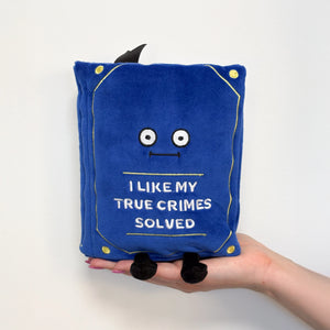 A blue plush book-shaped pillow with embroidered eyes, a stitched smile, gold corner accents, and the text 'I LIKE MY TRUE CRIMES SOLVED' on the front. A hand holds the pillow against a plain white background.