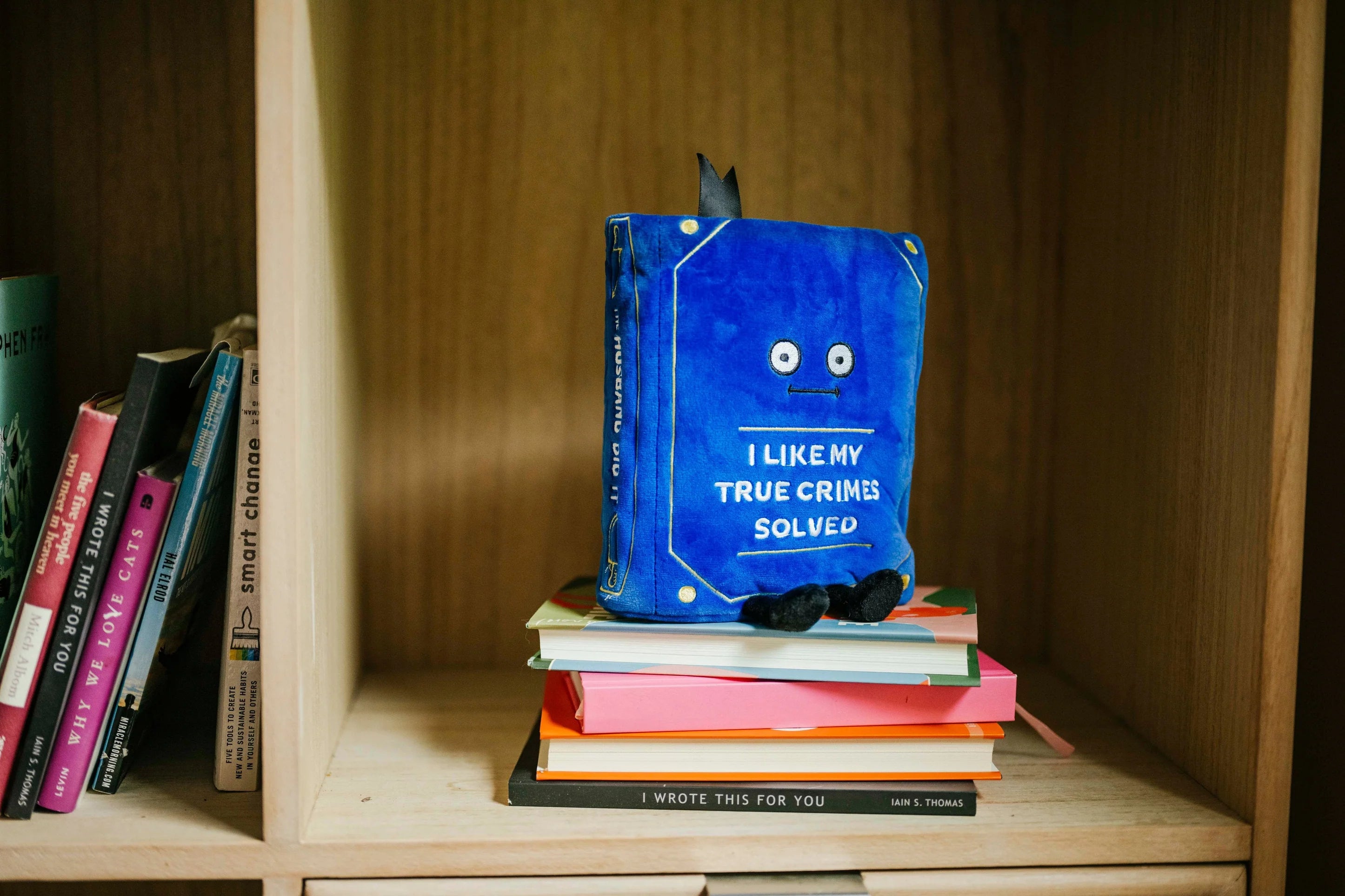 A blue hardcover book titled 'I LIKE MY TRUE CRIMES SOLVED' with cartoon eyes and a small black bow on top, displayed on a stack of colorful books inside a wooden bookshelf.