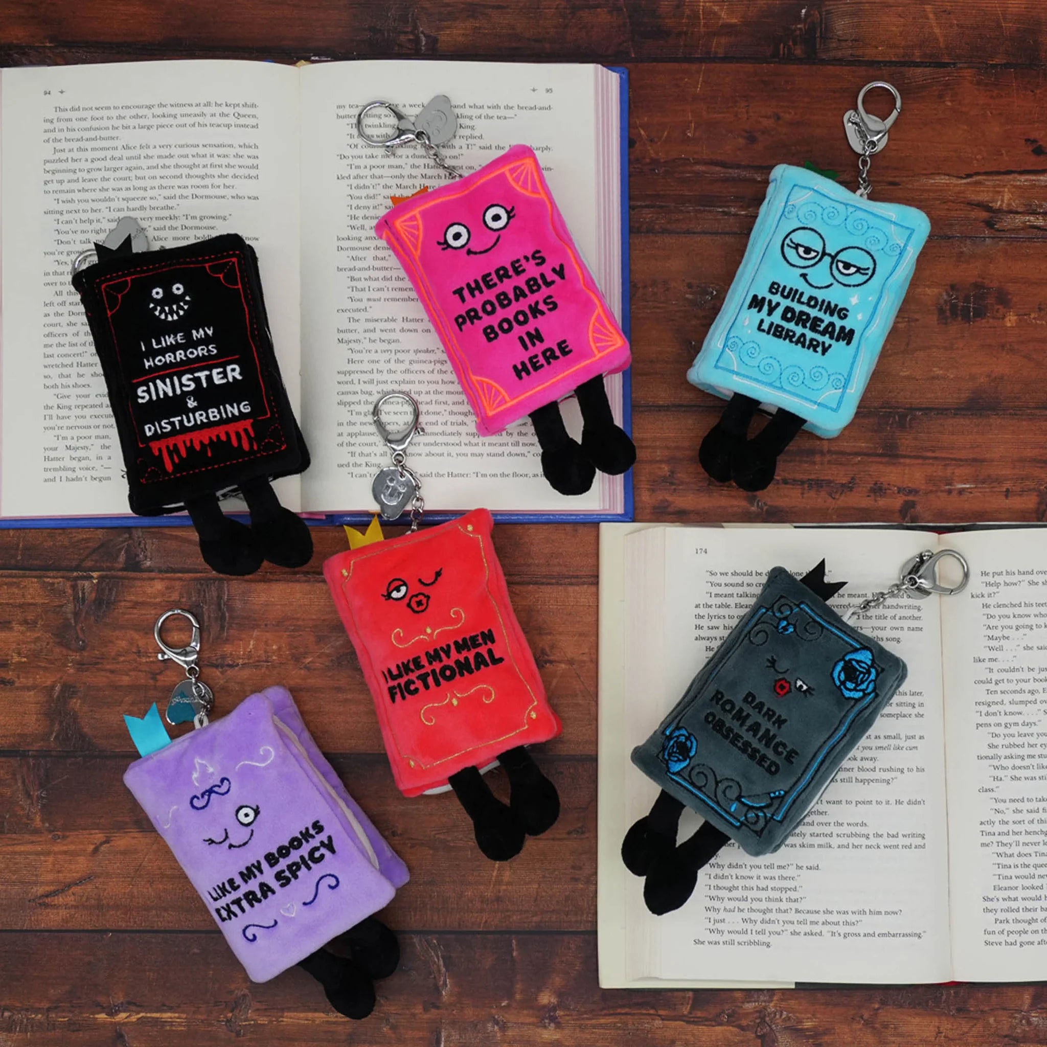 Five plush book-shaped keychains with black legs and metal keyrings are arranged on an open book over a wooden surface. The books are black, pink, light blue, purple, and red, each with humorous text like 'My Sinister Disturbing Book' and 'There's Probably Books In Here'.