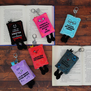 Five plush book-shaped keychains with black legs and metal keyrings are arranged on an open book over a wooden surface. The books are black, pink, light blue, purple, and red, each with humorous text like 'My Sinister Disturbing Book' and 'There's Probably Books In Here'.