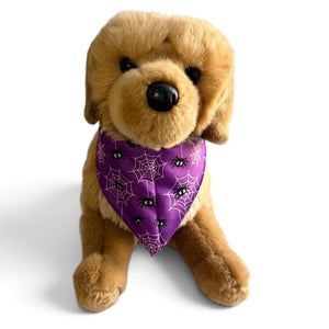 A golden retriever plush toy wearing a purple Halloween bandana with spiderwebs and black spider eyes, set against a plain white background.