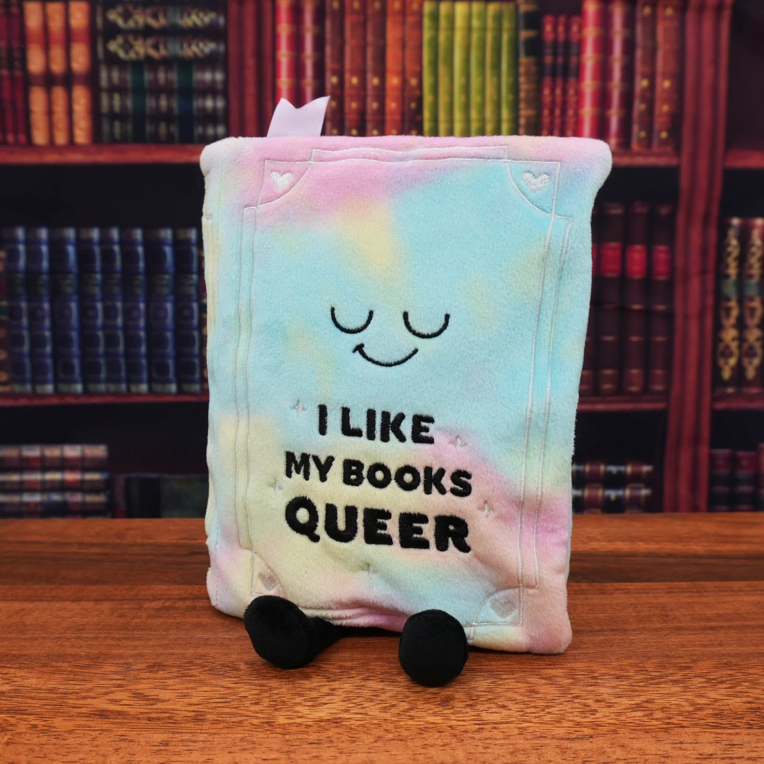 A plush book character toy with pastel tie-dye colors, featuring a sleeping face and the text 'I LIKE MY BOOKS QUEER' on the front, standing on a wooden table with a blurred bookshelf background.