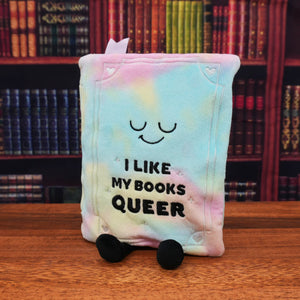 A plush book character toy with pastel tie-dye colors, featuring a sleeping face and the text 'I LIKE MY BOOKS QUEER' on the front, standing on a wooden table with a blurred bookshelf background.