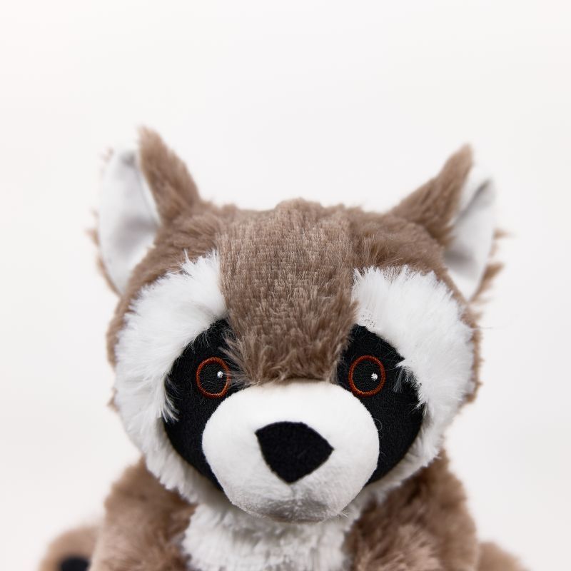 A close-up of a plush raccoon toy against a plain white background. The raccoon has brown and white fur with black around its eyes and a white snout, and small orange-rimmed eyes.