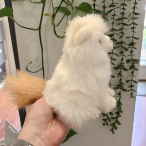 White fluffy toy held in a hand with green plants in the background