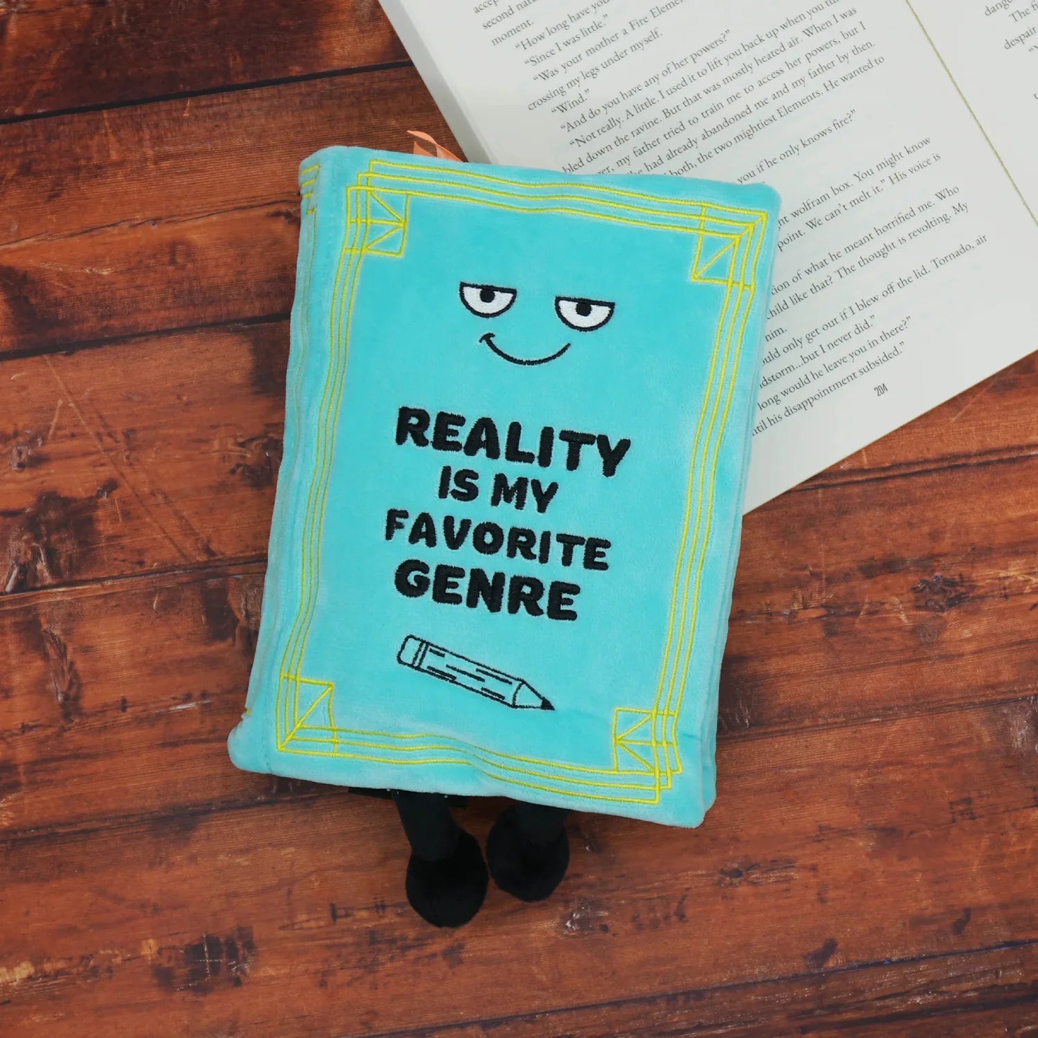 Blue pillow with 'Reality is my favorite genre' text on a wooden surface with an open book.