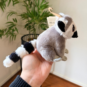 A small plush ring puppet of a ring-tailed lemur being held in a person's hand, with a potted plant and indoor room background visible.