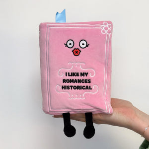 A pink plush book character being held by a hand. The plush has embroidered eyes, a red star-shaped mouth, the text 'I LIKE MY ROMANCES HISTORICAL' on the front, black legs, and a small blue ribbon bookmark at the top.