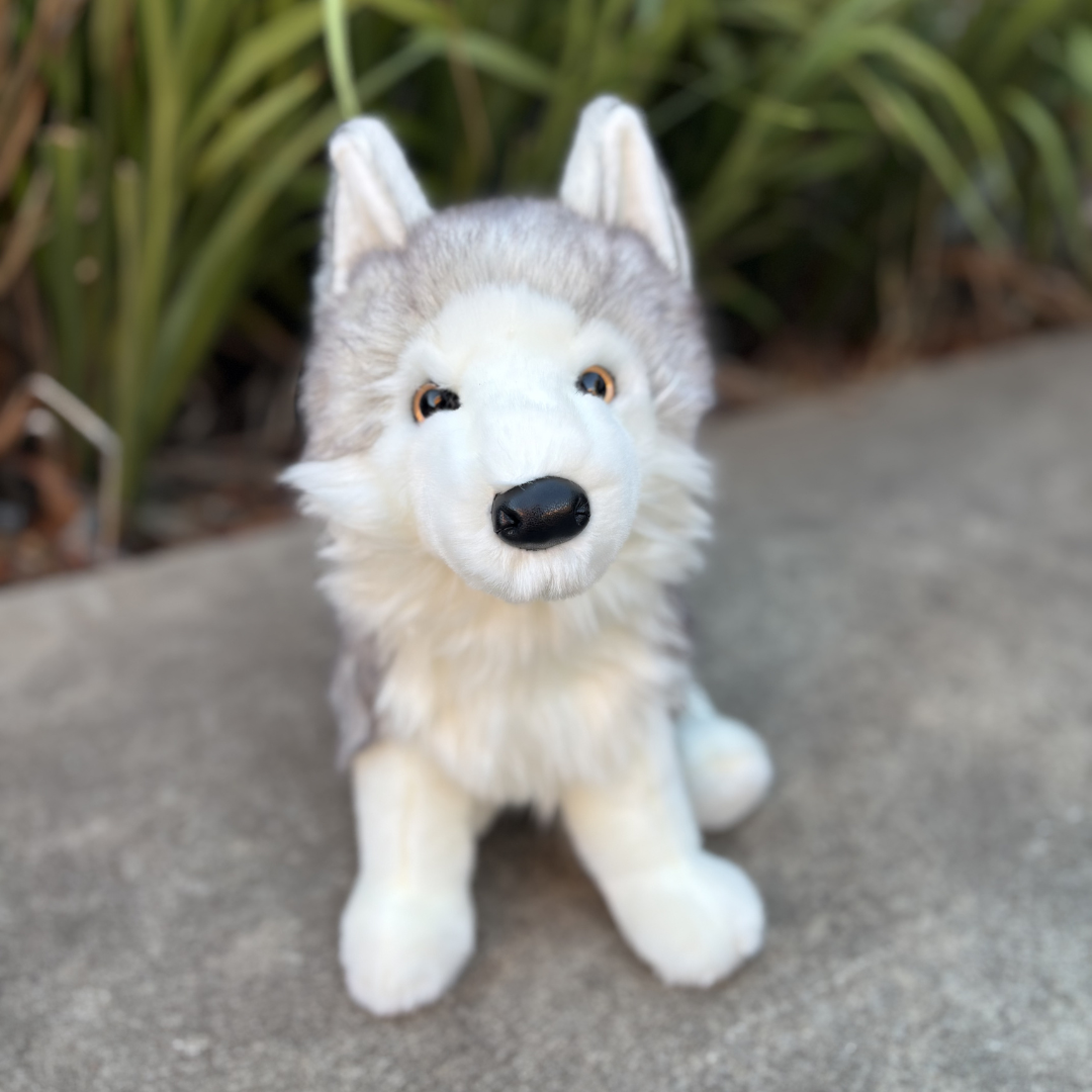Shadow White Wolf by Douglas Cuddle Toy sitting in front of green bushes