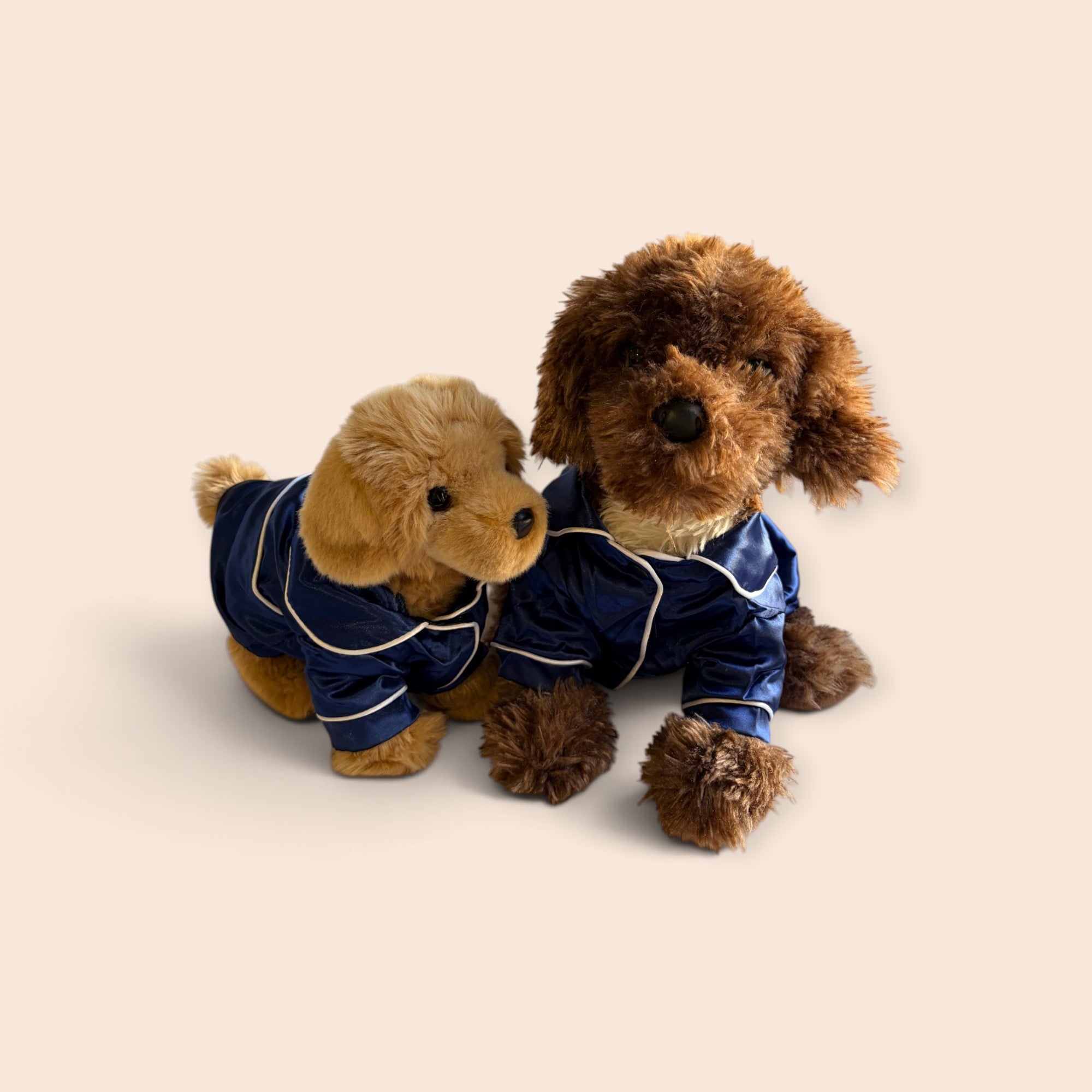 Two plush dog toys wearing matching navy blue pajamas with white piping, posed together on a plain warm beige background.