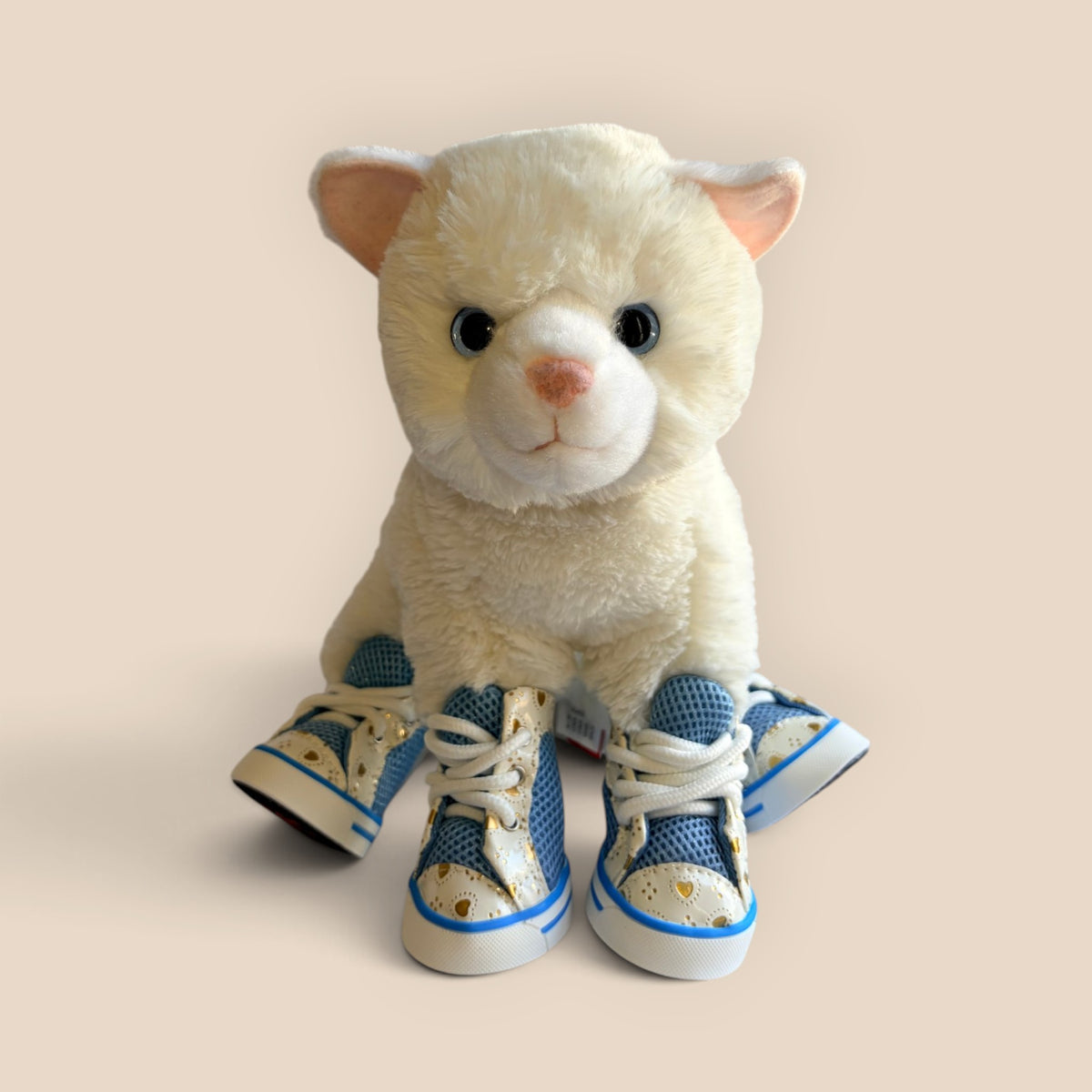 A white plush cat toy wearing blue and white high-top sneakers with gold heart details, posed against a solid light beige background.