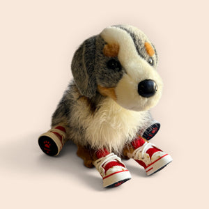 A plush dog toy with a gray, cream, and brown fur pattern, wearing red and white sneakers, set against a solid pale peach background.
