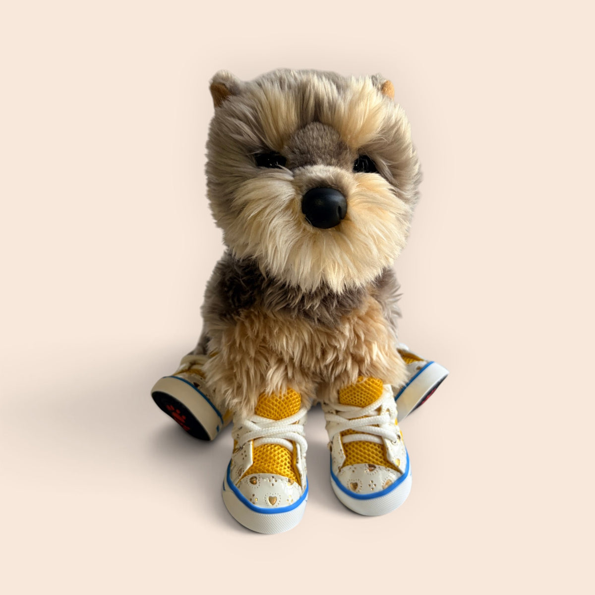 A plush dog toy with tan and brown fur, wearing yellow and white sneakers with blue trim, posed against a solid pale beige background.