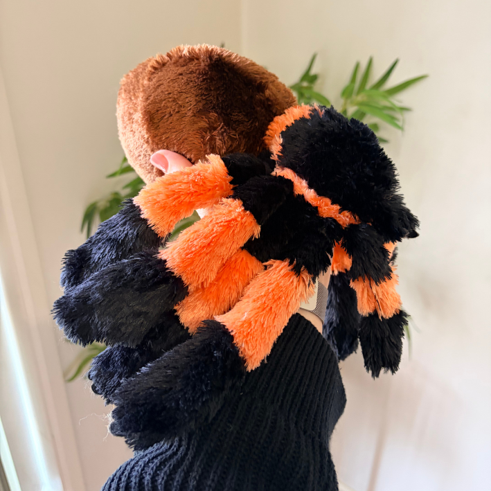 A person wearing a plush spider costume accessory with black and orange fuzzy legs and a brown furry head, photographed indoors against a light wall with a plant in the background.