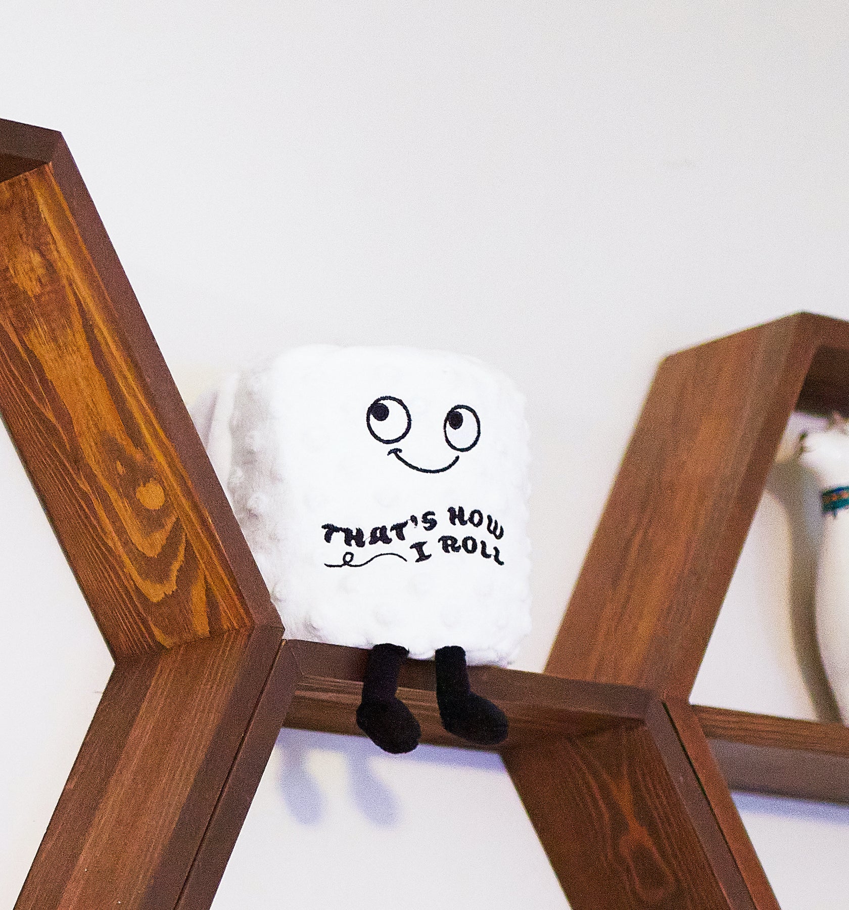 A white plush square pillow with a black embroidered face and the text 'THAT'S HOW I ROLL' sits on a wooden shelf against a white wall.