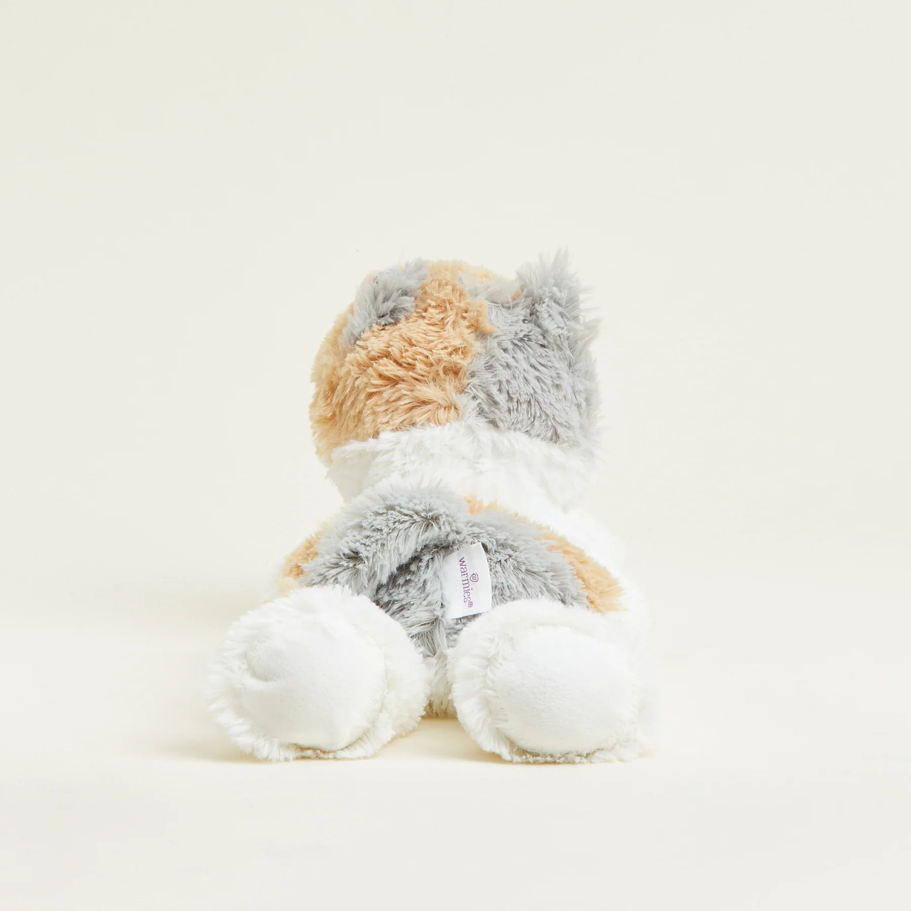 A plush stuffed animal toy shaped like a cat, photographed from the back against a clean, solid, warm off-white background. The cat features fluffy fur in white, light gray, and a soft peachy-orange color.