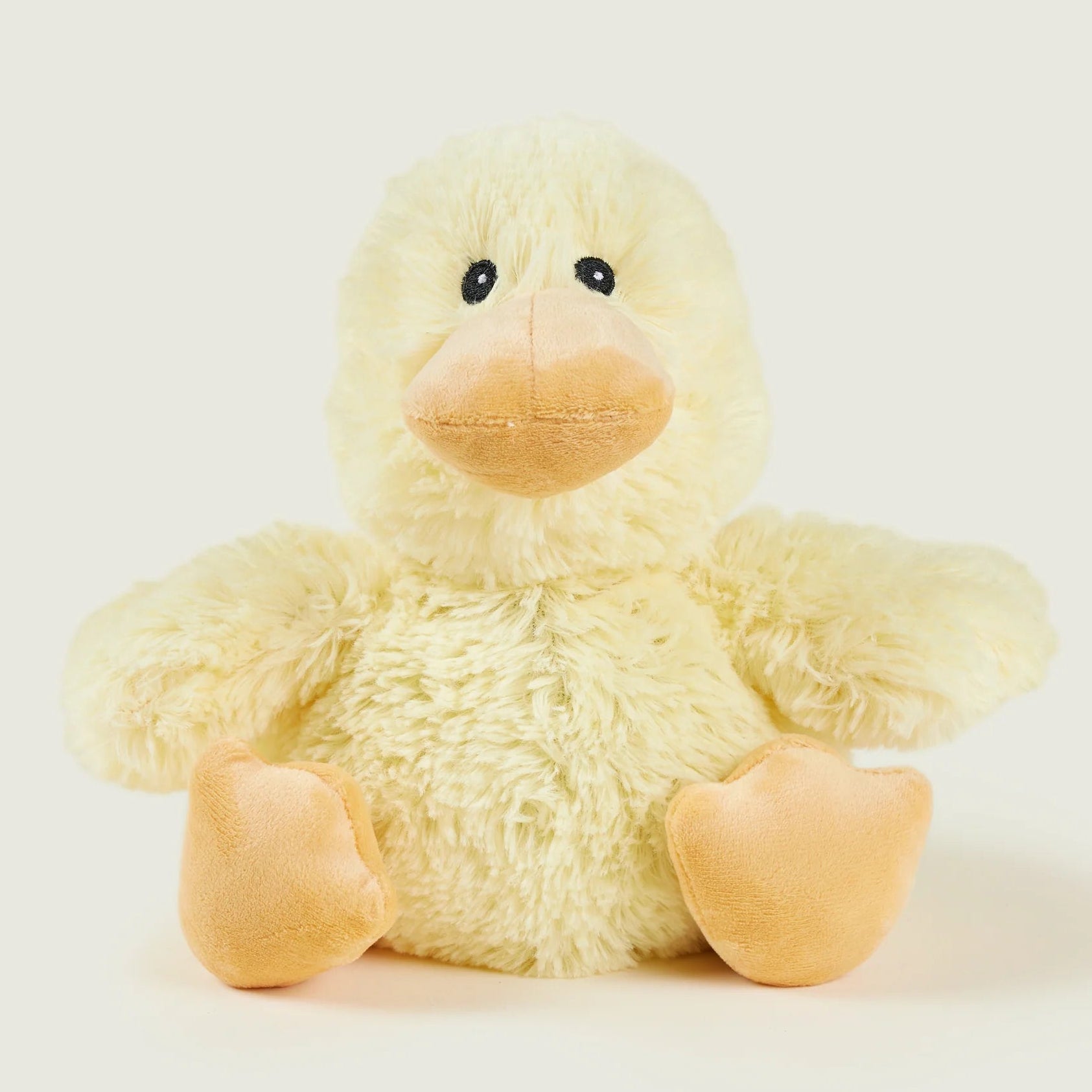 A small plush yellow duck toy with a soft fuzzy body, peach-colored beak and feet, and black bead eyes, photographed against a light warm gray background.