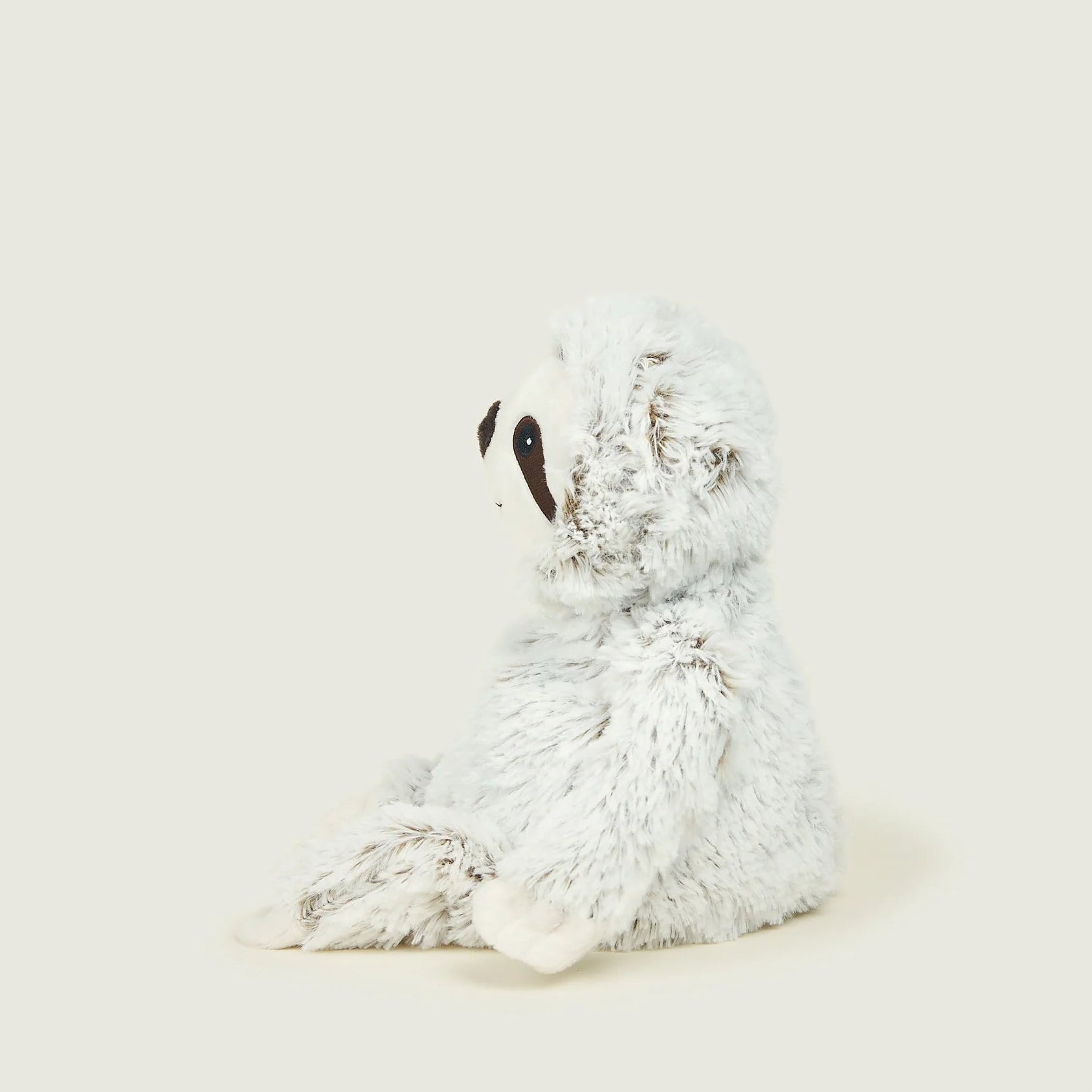 A plush sloth toy in white and light gray faux fur, sitting in profile against a solid, warm off-white background.