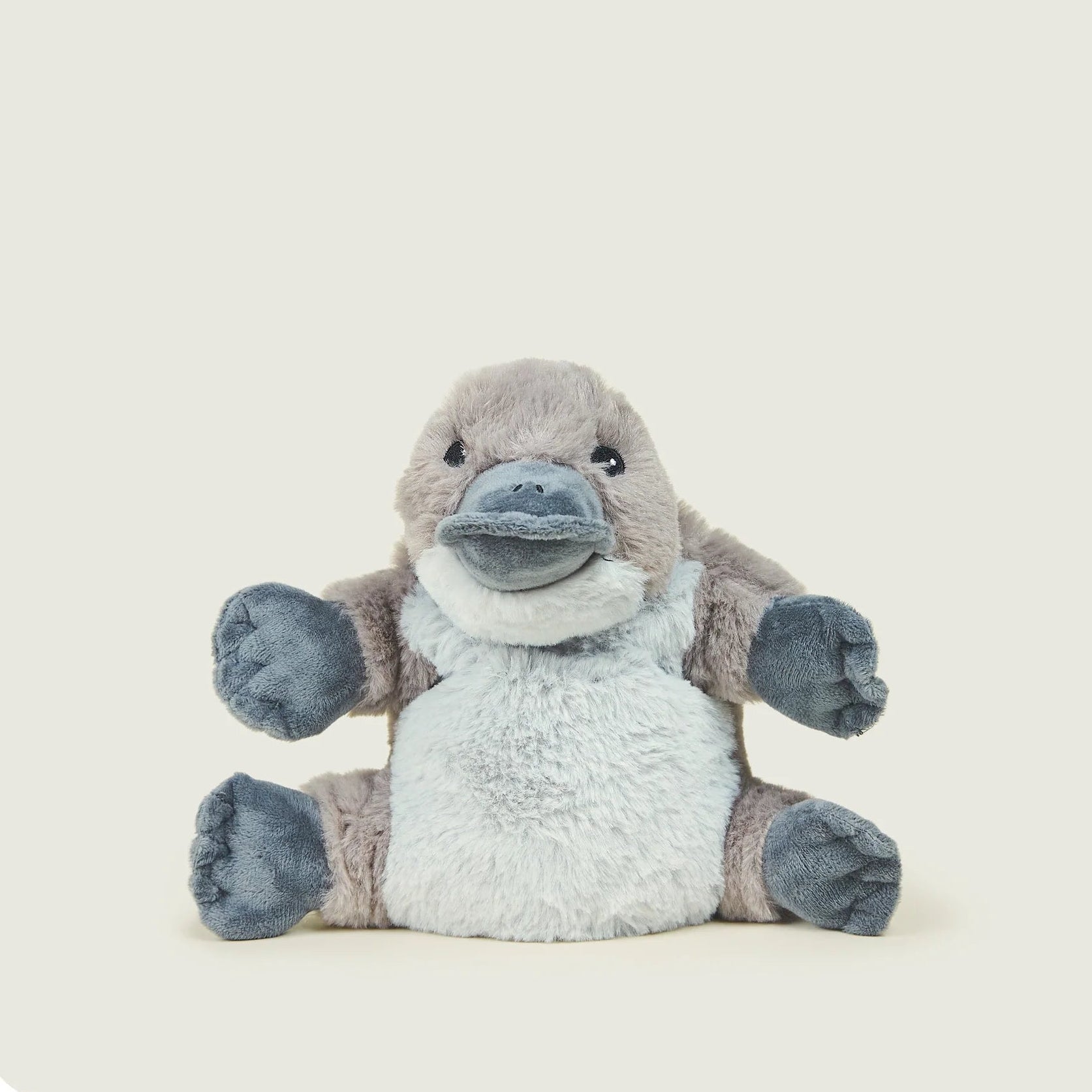 A plush toy of a sitting platypus, featuring a light gray fuzzy body, brownish-gray head and arms, and dark gray paws and beak, set against a solid cream-colored background with rounded corners.