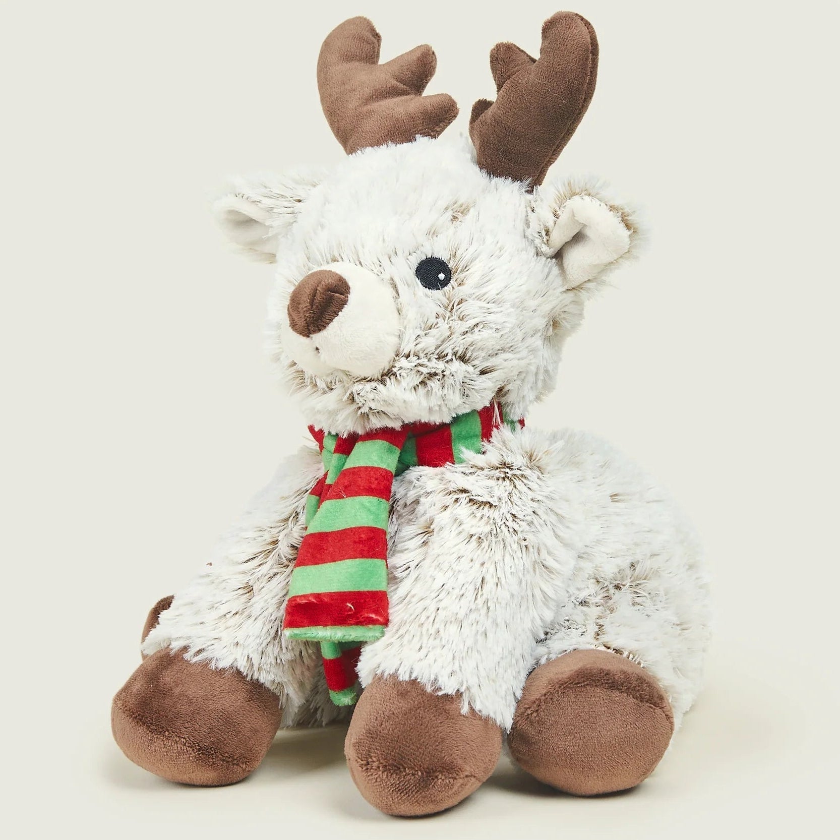 A white plush reindeer toy with brown antlers, hooves, and snout, wearing a red and green striped scarf, photographed against a light warm gray background.