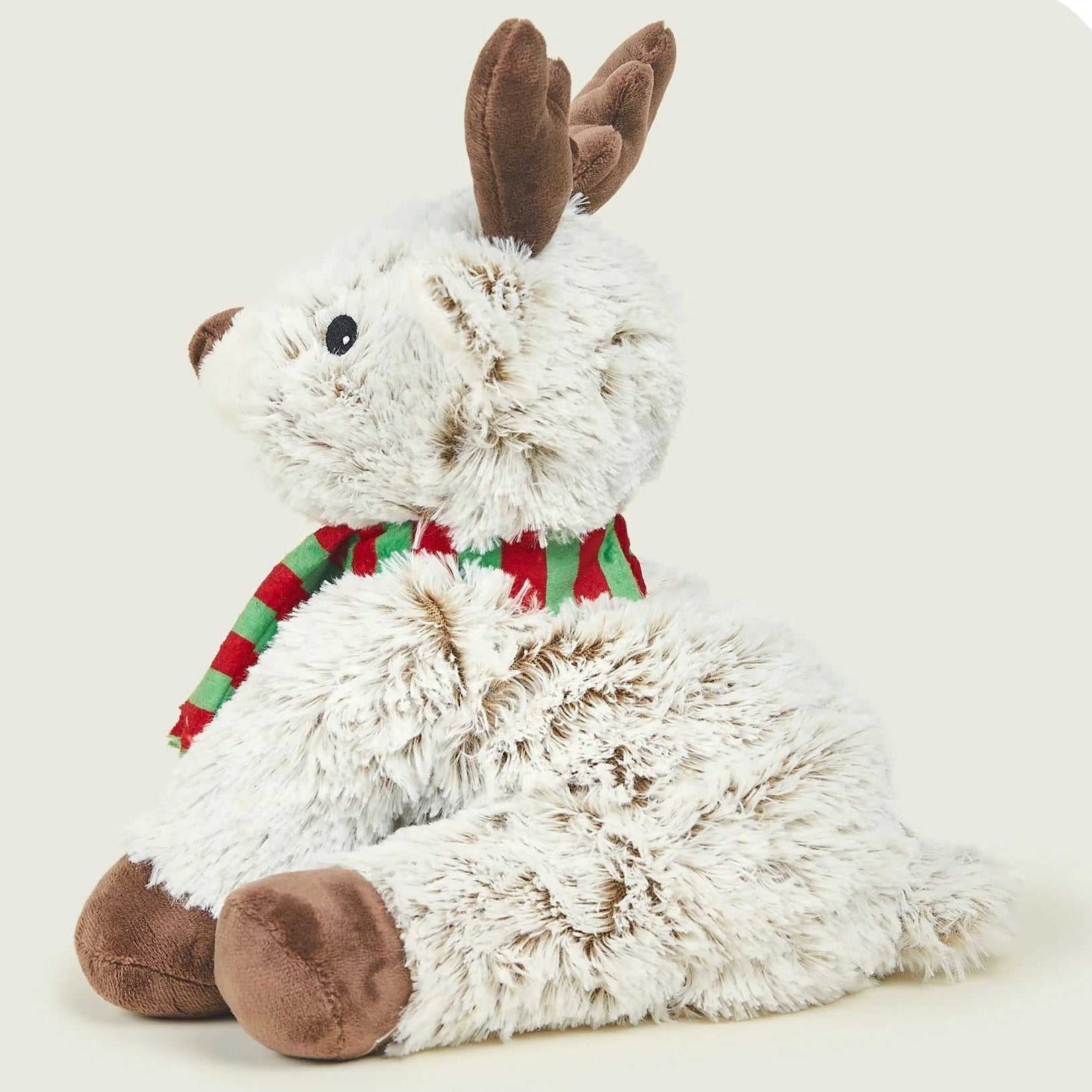 A plush stuffed animal shaped like a reindeer, with fluffy off-white fur, brown ears and hooves, and a red and green striped scarf, photographed on a light warm gray background.