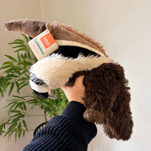 A plush badger toy being held by a person's hand, with a product tag reading 'MILD Cuddlerkins' attached. The background shows a light-colored wall and a green plant.
