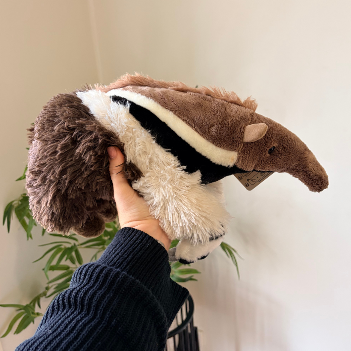 A plush animal toy being held up by a hand. The toy has a long brown snout, a black and white body, and fluffy brown and white fur.
