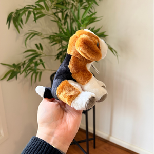 A small plush dog toy with tan, black, and white fur is being held in a person's hand in an indoor setting with a potted plant and wooden floor in the background.