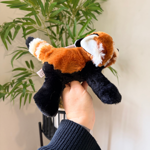 A plush red panda toy being held by a hand, with a potted plant and cream-colored wall in the background.