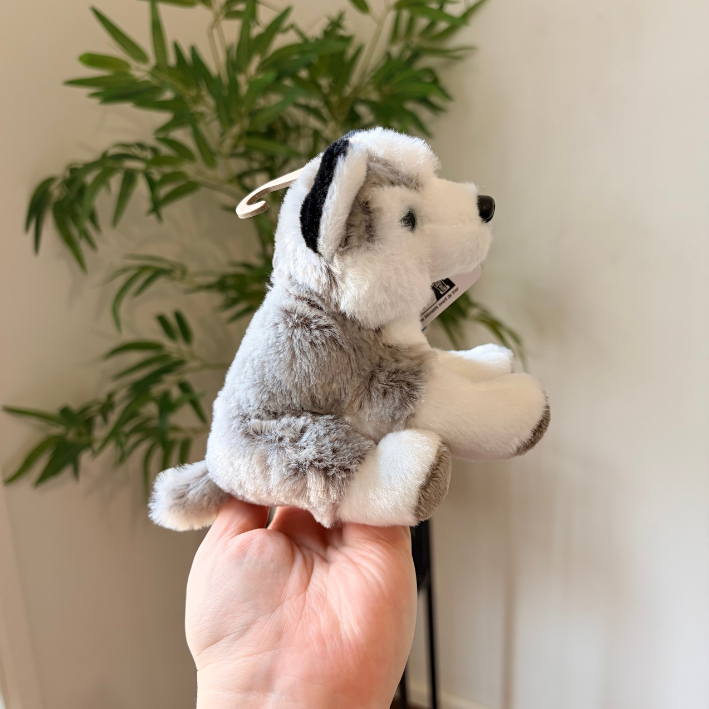 A small plush dog toy with gray, white, and black fur is being held in a person's hand in front of a potted green plant and a light-colored wall.