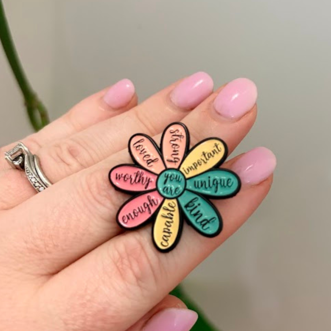 Colorful flower-shaped pin with motivational phrases held by a hand with pink nails.